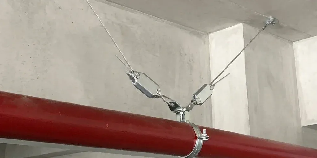 Acrefine ASB-GRP seismic cable bracing on a red sprinkler pipe: two wire-rope restraints with wire-rope grips, joined at the pipe clamp